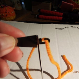 May include: A close-up of a hand holding a needle threader with orange yarn threaded through it. The needle threader is on a white surface with a black thread and a date, 3/30/2020, written on it.