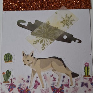 May include: A white card featuring a copper glitter top strip, a metallic silver tool secured with snowflake-patterned tape, a central coyote illustration, and decorative stickers of two cacti and a bottom border of colorful butterflies.