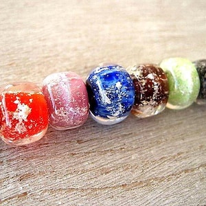 May include: A close-up of a strand of colorful glass beads. The beads are in various colors, including teal, red, pink, blue, brown, green, black, and purple. Each bead has a unique pattern and texture, with a speckled or marbled appearance.