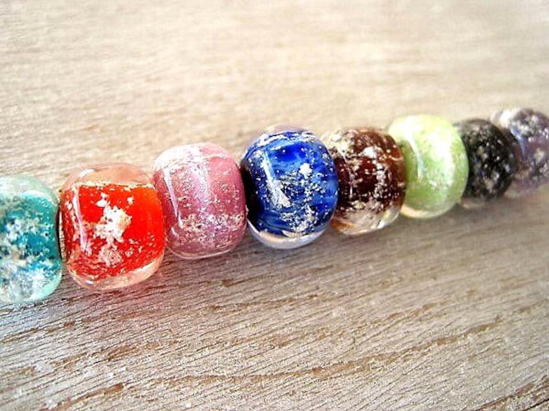 May include: A close-up of a strand of colorful glass beads. The beads are in various colors, including teal, red, pink, blue, brown, green, black, and purple. Each bead has a unique pattern and texture, with a speckled or marbled appearance.