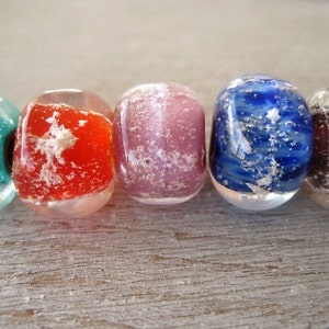 May include: Close-up of five colorful, oval-shaped glass beads. The beads are in shades of teal, red, pink, blue, and brown, with a textured, cloudy interior. The beads are displayed on a light-colored wooden surface.