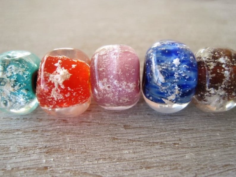 May include: Close-up of five colorful, oval-shaped glass beads. The beads are in shades of teal, red, pink, blue, and brown, with a textured, cloudy interior. The beads are displayed on a light-colored wooden surface.
