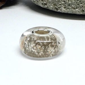 Pode incluir: Uma conta redonda e transparente com um núcleo prateado. O interior da conta é preenchido com partículas castanhas claras e brancas, criando uma aparência texturizada. A conta está sobre um fundo branco.