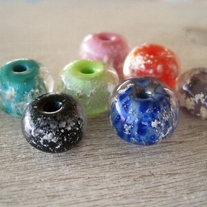 May include: A collection of colorful, round glass beads with a hole through the center. The beads are in various colors, including teal, pink, red, green, blue, black, and purple. Each bead has a speckled, frosted appearance.