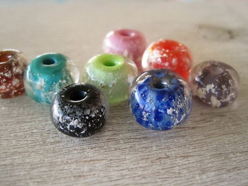 May include: A collection of colorful, round glass beads with a hole through the center. The beads are in various colors, including teal, pink, red, green, blue, black, and purple. Each bead has a speckled, frosted appearance.