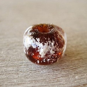 May include: A close-up of a clear, amber-colored bead with a hole through the center. The bead has white, frosted-looking inclusions throughout. The bead is sitting on a light brown, wooden surface.