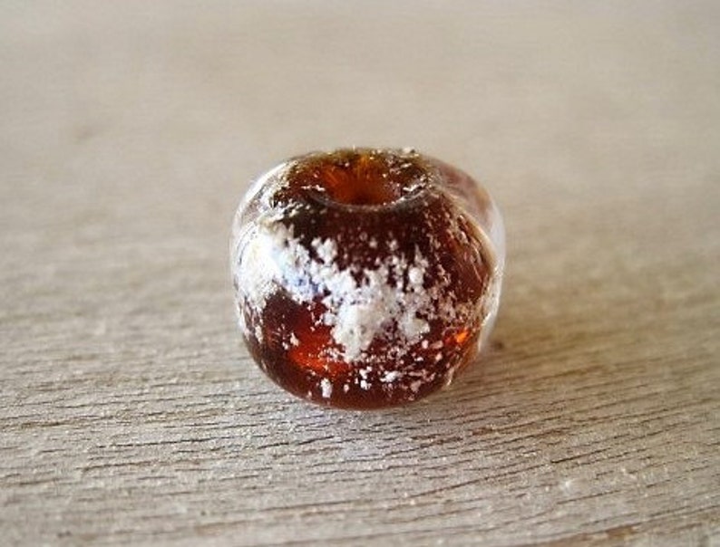 May include: A close-up of a clear, amber-colored bead with a hole through the center. The bead has white, frosted-looking inclusions throughout. The bead is sitting on a light brown, wooden surface.
