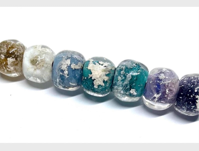 May include: A strand of colorful, translucent beads. The beads are a mix of colors, including brown, white, blue, teal, purple, and dark blue. Each bead has a unique, textured appearance, with variations in color and opacity. The beads are strung together on a white background.