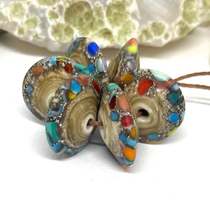 May include: A cluster of round, disc-shaped beads strung on a thin brown cord. Each bead features a central tan, wood-like swirl pattern surrounded by a textured outer rim embedded with small, multi-coloured fragments in red, blue, orange, and yellow.