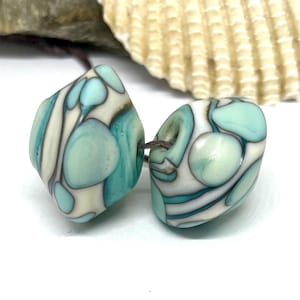 May include: Two teal and white swirl glass beads with a white background.
