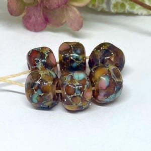 May include: Six colorful artisan beads strung on a beige cord. The beads feature a mosaic pattern with shades of brown, red, blue, and pink. The beads are irregularly shaped and have a glossy finish, perfect for jewelry making.