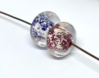 Op de afbeelding: Twee glazen kralen met een transparante buitenlaag en een gevlekte binnenlaag. Eén kraal is blauw en wit, de andere is rood en wit.