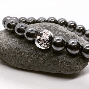 May include: A black hematite beaded bracelet with a clear glass bead containing white and black flecks. The bracelet is resting on a gray stone.