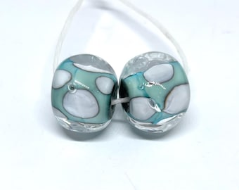 May include: Two glass beads with a teal and white speckled pattern. The beads are round and have a clear glass finish.