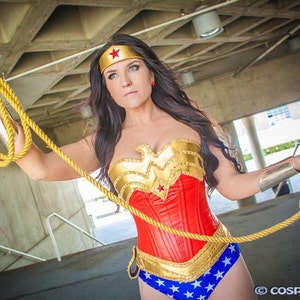 May include: A woman in a Wonder Woman costume, featuring a red corset with gold accents, blue star-spangled shorts, and a gold headband with a red star. She holds a golden rope.