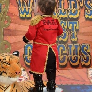 May include: A young child dressed as a ringmaster stands on a barrel in front of a circus backdrop. The child is wearing a red and gold costume with the text "PAL The Magnificent" on the back. A tiger plush toy is in the foreground.