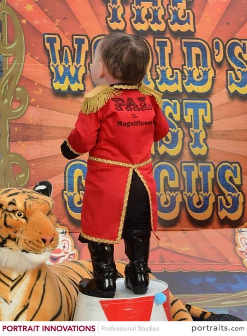 May include: A young child dressed as a ringmaster stands on a barrel in front of a circus backdrop. The child is wearing a red and gold costume with the text "PAL The Magnificent" on the back. A tiger plush toy is in the foreground.