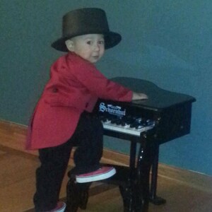 May include: A young child wearing a red jacket and a black top hat plays a black toy piano. The piano has the word "Schroeder" printed on it.