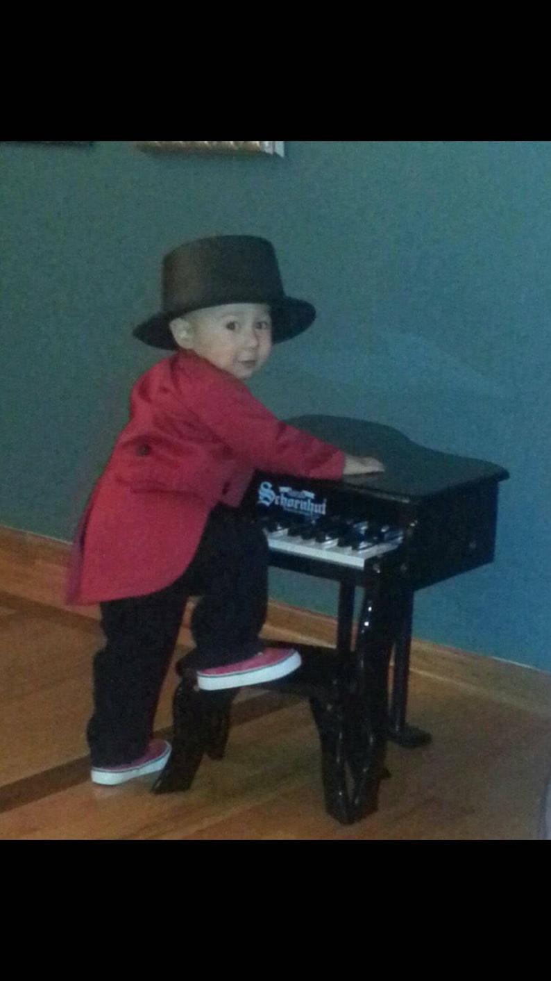May include: A young child wearing a red jacket and a black top hat plays a black toy piano. The piano has the word "Schroeder" printed on it.