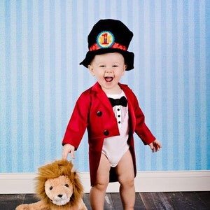 May include: A young child dressed as a ringmaster, wearing a red jacket with a tailcoat, a black top hat with a yellow and red number one on it, and a black bow tie. The child is holding a brown plush lion with a toothy grin.