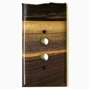 May include: A rectangular wooden light switch cover with a layered design. The top section is dark brown, transitioning to lighter brown and tan hues. Two white toggle switches and two small dark brown screw holes are visible.