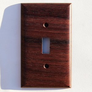 May include: A rectangular wooden light switch cover in a dark brown hue. It features a rectangular aperture for the switch and two small circular holes. The wood grain is visible, providing a textured appearance to the design.