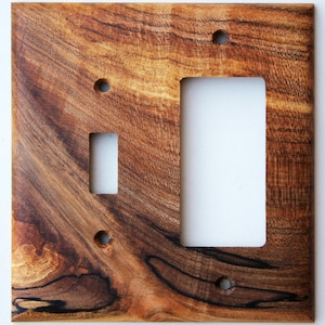 May include: A wooden light switch cover with a natural wood grain pattern. The cover has openings for a single-pole switch and a standard outlet, along with screw holes for mounting. The wood has a warm, brown tone.