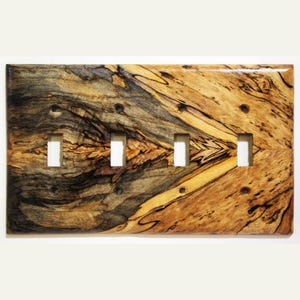 Könnte beinhalten: Eine Holz-Lichtschalterplatte mit vier rechteckigen Öffnungen. Die Platte hat ein natürliches Holzmaserungsmuster mit Schattierungen von Braun, Schwarz und Beige. Die Holzmaserung hat ein einzigartiges, wirbelndes Muster. Die Platte ist rechteckig und hat eine glänzende Oberfläche.