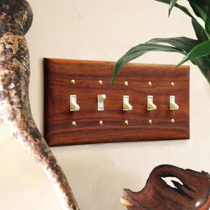 Könnte beinhalten: Eine Holz-Wandplatte mit fünf Lichtschaltern und Messing-Schrauben. Die Platte ist in einem satten Rotbraun gehalten und hat eine Holzmaserung.