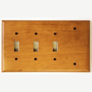 Peut inclure: Une plaque d'interrupteur en bois avec trois ouvertures pour interrupteurs à bascule et six trous de vis. La plaque est de couleur brun moyen chaud avec une finition brillante. Le grain du bois est visible.
