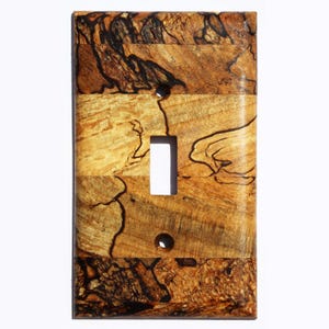 May include: A light switch cover made of wood with a natural wood grain pattern. The cover is a light brown color with dark brown streaks throughout. The switch cover is rectangular and has a single switch opening.