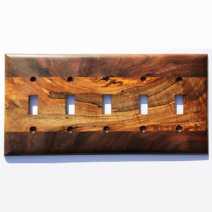 May include: A six-gang wall plate made of dark brown wood with a lighter brown wood inlay. The plate has six rectangular cutouts for light switches.