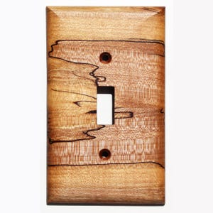 May include: A light brown wooden light switch cover with a natural wood grain pattern and dark brown streaks. The cover has a single switch opening and two screw holes.