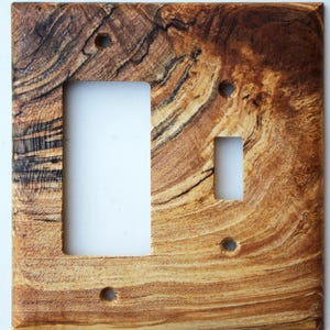 May include: Wooden light switch cover with a natural wood grain pattern. The cover has openings for a standard light switch and an outlet, with screw holes in each corner. The wood features a mix of light and dark brown tones.