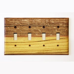 Könnte beinhalten: Eine Holz-Lichtschalterplatte mit vier Kippschaltern. Die Platte besteht aus zweifarbigem Holz, mit einem hellgelb-braunen Farbton unten und einem dunkleren Braunton oben. Die Schalter sind weiß und die Schraubenlöcher sind schwarz.