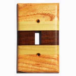May include: A wooden light switch cover with a multi-striped design. The cover is made of three different types of wood: a light wood, a dark wood, and a medium-toned wood. The cover has a rectangular shape and a single switch opening.