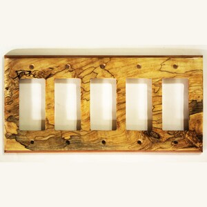 Puede incluir: Una placa de interruptor de luz de madera con cinco aberturas rectangulares. La placa está hecha de madera con un patrón de vetas natural en tonos marrón, beige y negro. La placa tiene forma rectangular con esquinas redondeadas.