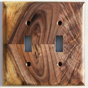 May include: A square, wooden double light switch cover with a dark brown wood grain pattern. It features two rectangular switch openings and four screw holes. The wood grain has a swirling pattern, giving it a natural appearance. The cover is made of wood.