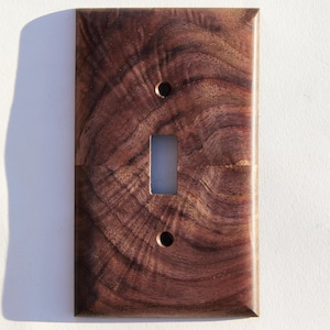 May include: A rectangular, wooden light switch cover with a single toggle opening and two screw holes. The cover is made of dark brown wood with a swirling grain pattern. The wood has a natural, polished finish.