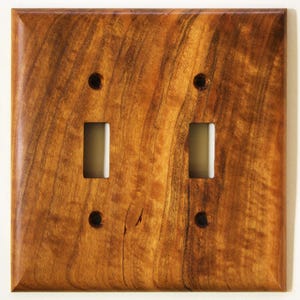 Peut inclure: Plaque de recouvrement double pour interrupteur en bois avec un fini grain de bois naturel. La plaque a deux ouvertures rectangulaires pour les interrupteurs et quatre petits trous pour les vis de fixation. Le bois est brun.