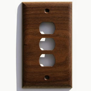 May include: A rectangular, wooden wall plate with a dark brown finish. It has three outlet openings and two screw holes. The plate is designed for electrical outlets. The dimensions are in centimetres.