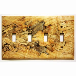 Könnte beinhalten: Eine Holz-Lichtschalterabdeckung mit vier rechteckigen Öffnungen für die Schalter. Die Abdeckung hat ein natürliches Holzmaserungsmuster mit Farbtönen von Beige, Braun und Schwarz. Die Holzmaserung hat ein einzigartiges, wirbelndes Muster.