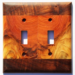 May include: A double toggle light switch cover made of wood with a natural wood grain pattern. The cover is made of two different types of wood, one with a darker brown color and the other with a lighter orange color. The cover has a rectangular shape and is designed to fit over a standard double toggle light switch.