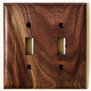 Può includere: Piastra di copertura per interruttore a doppia luce in legno quadrato. La piastra è realizzata in legno marrone scuro con venature visibili. Ha due aperture rettangolari per interruttori e quattro fori per viti. I bordi sono leggermente smussati.
