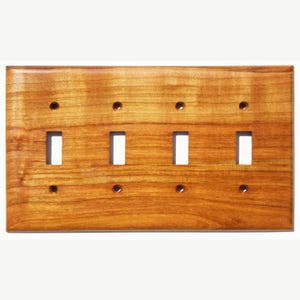 Könnte beinhalten: Eine rechteckige Lichtschalterplatte aus Holz mit vier Schalteröffnungen und acht Schraubenlöchern. Die Platte hat eine warme, rotbraune Oberfläche mit sichtbarer Holzmaserung.
