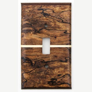 Puede incluir: Una placa de pared rectangular para interruptor de luz con un patrón de veta de madera marrón y marcas orgánicas oscuras y arremolinadas. Una fina franja incrustada de color crema cruza horizontalmente el centro de la placa.