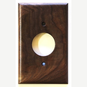 Pode incluir: Uma placa de parede retangular de madeira castanha escura com uma grande abertura circular e dois orifícios menores. A placa tem um acabamento liso e polido, e o grão da madeira é visível. A abertura circular é para uma tomada elétrica.