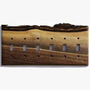 May include: A wooden light switch plate with six switch openings. The plate is made of dark brown wood with lighter brown and black streaks. The top edge has a rough, natural bark edge.