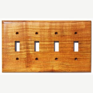 Könnte beinhalten: Eine rechteckige Lichtschalterplatte aus Holz mit vier Kippschalteröffnungen und vier Schraubenlöchern. Die Platte hat eine warme, mittelbraune Farbe mit sichtbarer Holzmaserung.