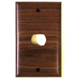 May include: A rectangular, dark brown wooden light switch cover with a single, circular opening. The wood grain is clearly visible. Two small, black screws are positioned above and below the opening. The edges of the cover are slightly rounded.